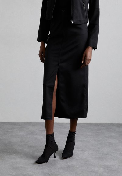 Zwarte midi-rok met een voorkap, gecombineerd met strakke zwarte enkellaarsjes. De outfit heeft een elegante, minimalistische uitstraling en een gladde textuur.