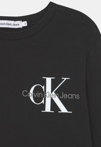 Maglietta nera in cotone a maniche lunghe con scollo rotondo e logo bianco "CK" con la scritta "Calvin Klein Jeans" sottostante. Design semplice e moderno.