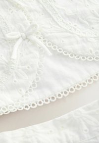 Tissu blanc avec motifs floraux brodés, bords festonnés et un petit nœud noué sur un matériau léger.