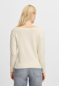 Maglione color crema chiaro con scollatura ampia, polsini e orlo a coste. Tessuto morbido, vestibilità leggermente rilassata, abbinato a jeans grigi chiari.