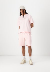 Helles rosa Baumwoll-T-Shirt mit kurzen Ärmeln, passende rosa Shorts und eine beigen Mütze. Das Shirt hat ein einfaches Logo und dazu lässige Sneakers.