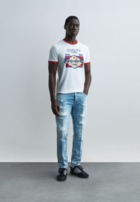 Jeune homme debout, main dans la poche, portant un t-shirt blanc à motifs avec bordure rouge, un jean bleu clair déchiré et des baskets noires.