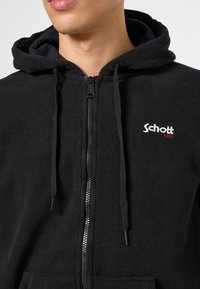 Schwarzer Fleece-Kapuzenpullover mit durchgehendem Reißverschluss, Kordelzug am Kapuzen, und besticktem "Schott"-Logo auf der linken Brust. Weiche Textur und lässiges Design.