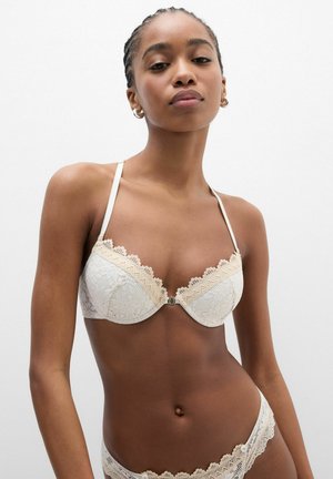 Mujer con sujetador de encaje blanco y ropa interior a juego, de pie frente a un fondo blanco liso.