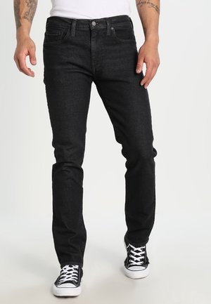Mand iført sorte slim-fit jeans, hvid skjorte, sort-hvide sneakers, med tatoverede underarme synlige mod en hvid baggrund.
