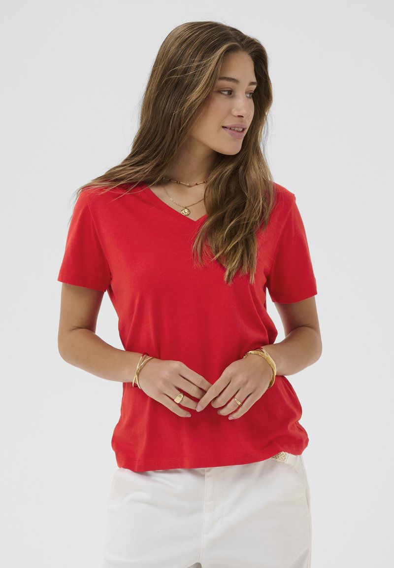 Giovane donna con lunghi capelli castani che indossa una maglietta rossa a scollo a V, pantaloni bianchi e gioielli dorati, che guarda verso destra.