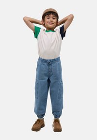 Enfant portant une chemise à manches courtes blanche et verte, un pantalon cargo bleu avec des poignets élastiques, et des chaussures à lacets brunes, souriant avec les mains derrière la tête.