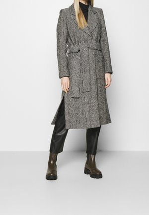 Personne portant un manteau long ceinturé à motif chevrons, un col roulé noir, un pantalon en cuir noir et des bottes épaisses marron sur un fond uni.