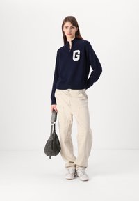 Marinblå zip-up tröja med vitt "G"-logotyp, kombinerad med ljusbeige, löst sittande byxor och en grå väska, med en mjuk textur och avslappnad stil.