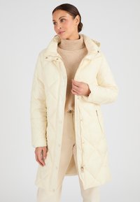 Giacca imbottita trapuntata color crema, con colletto alto, bottoni a pressione e zip laterali. Texture liscia con motivi a diamante per l'isolamento.