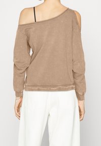 Sudadera marrón con escote de hombros descubiertos, mangas largas, textura suave, borde y puños acanalados, combinada con pantalones blancos.