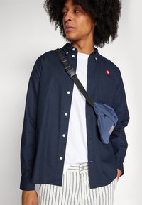 Navy button-up shirt with white buttons, worn over a white T-shirt. A small red logo on the chest and a blue utility bag with strap.