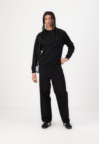 Schwarzes Crewneck-Sweatshirt in lässiger Passform, kombiniert mit lockeren schwarzen Hosen und schwarzen Sneakers mit weißen Akzenten. Kleine hellblaue Tasche umgehängt.