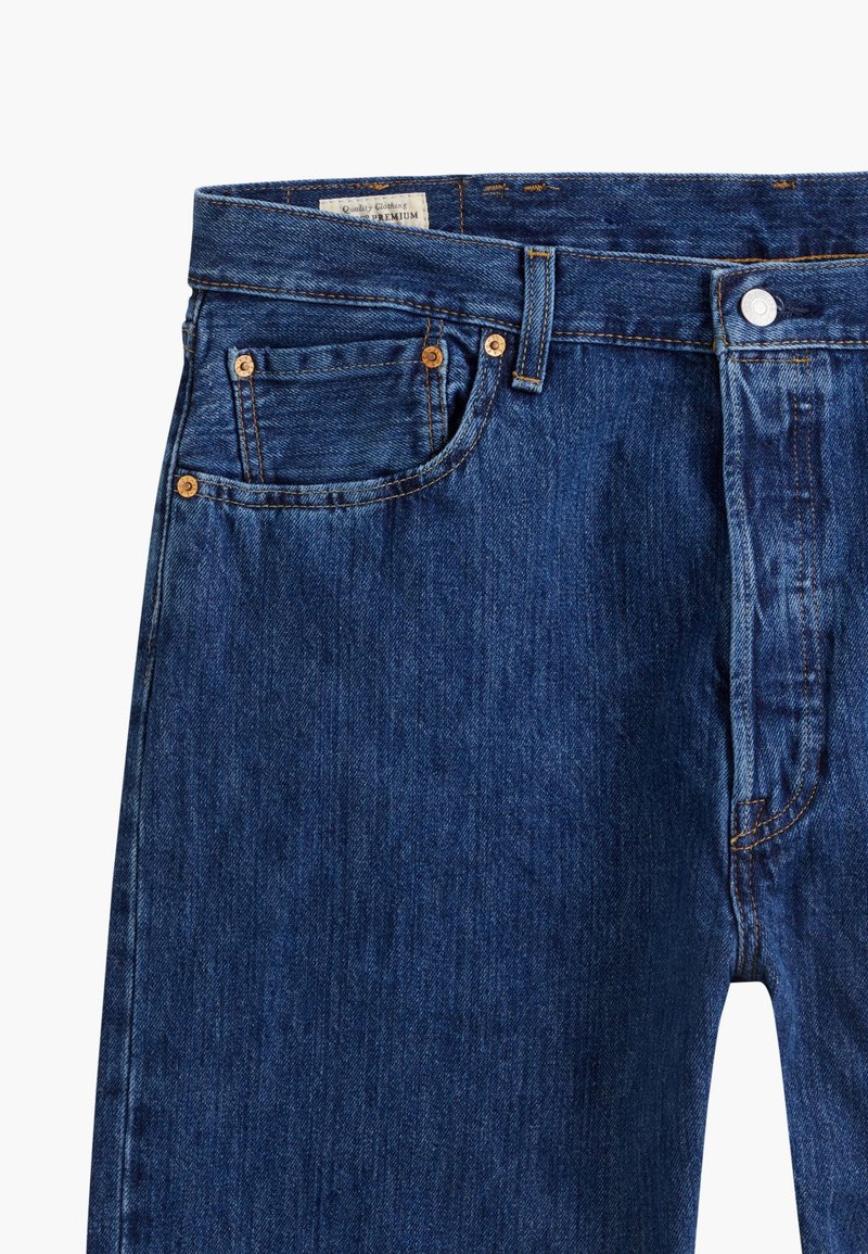 Jean en denim bleu avec des coutures dorées, design à cinq poches, fermeture à bouton en métal et une texture légèrement décolorée à la taille avant.