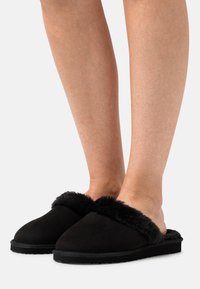 Svarte slip-on tøfler med kunstpelsdetaljer. Mykt semsket skinn på oversiden, polstret såle, rund tå og teksturert gummisåle for grep.