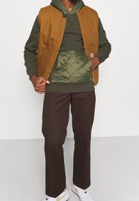 Homme portant un sweat à capuche matelassé vert olive avec poche avant, un gilet zippé marron, un pantalon marron foncé et des baskets blanches avec un swoosh beige.