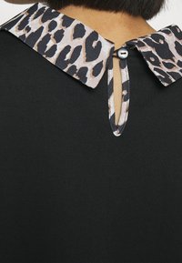 Svart topp med leopardmönstrad krage som är fäst med en svart knappöglan bakom en persons nacke, kort mörkt hår synligt.