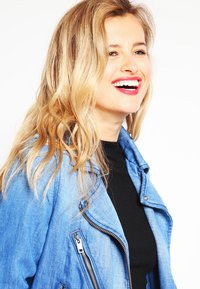 Giubbotto di jeans con risvolto aperto, dotato di zip argento e di un colore blu chiaro. Indossato sopra un top nero a coste.