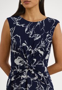 Marineblå kjole med hvitt blomstertrykk, korte ermer, og en knutedetalj i midjen. Mykt, teksturert stoff som faller elegant.