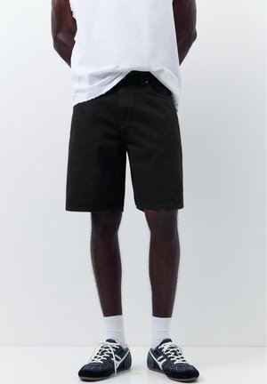 Mężczyzna w czarnych dżinsowych szortach, białych skarpetkach do kostek, czarno-białych sportowych butach i białej koszulce bez rękawów na jednolitym białym tle.