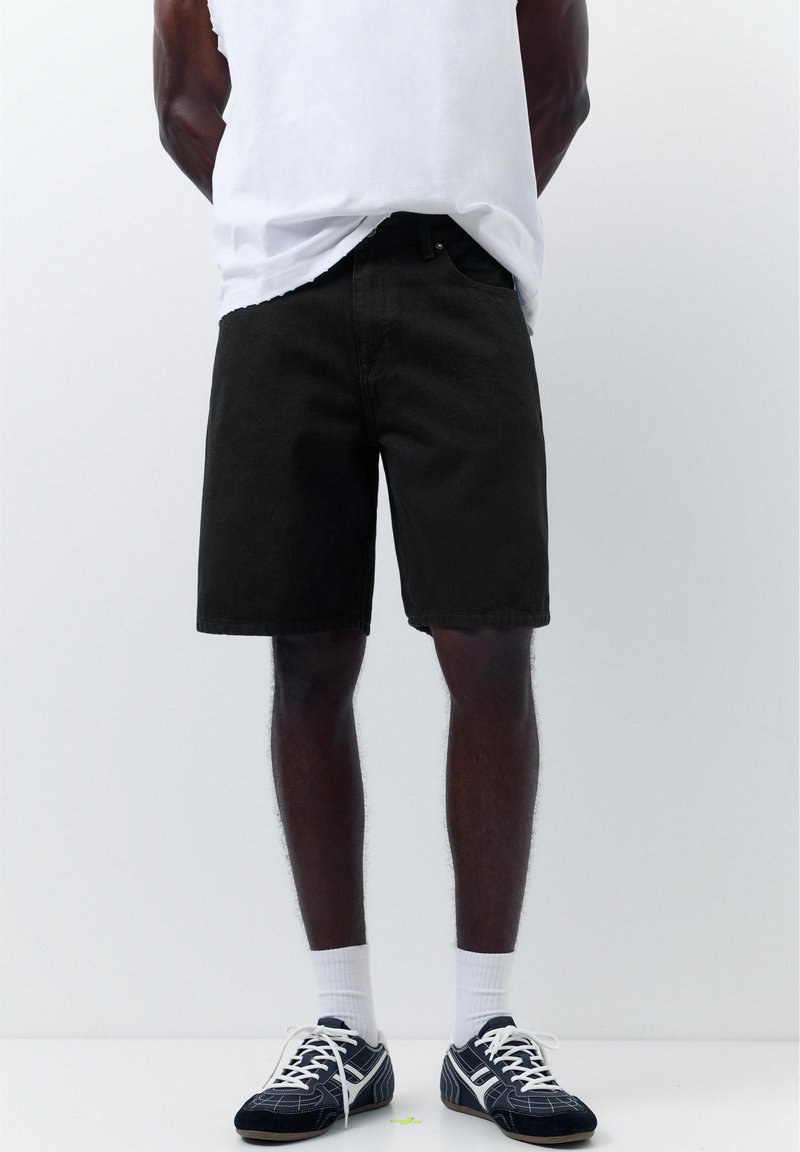 Homme portant un short en jean noir, des chaussettes blanches, des baskets noir et blanc et un débardeur blanc sur un fond blanc uni.