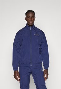 Marineblå vindjakke med zip-forside, høy krage og elastiske mansjetter. Har Sergio Tacchini-logoen på brystet.