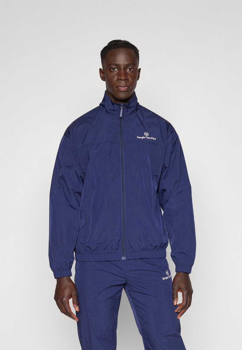 Marineblå vindjakke med zip-forside, høy krage og elastiske mansjetter. Har Sergio Tacchini-logoen på brystet.
