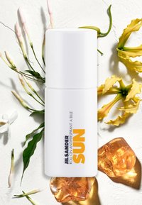 Vitt roll-on antiperspirant med en gul etikett som säger "SUN," mot en texturerad vit bakgrund med mjuka blommor och gelé-accenter.