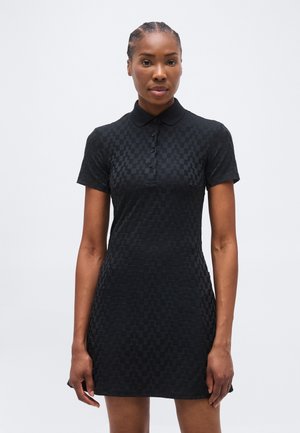 Femme aux cheveux tressés portant une robe polo à manches courtes noire à carreaux, debout devant un fond blanc uni.