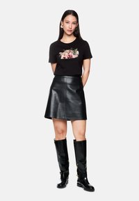 Schwarzes Baumwoll-T-Shirt mit Blumen- und Tierstickerei, kombiniert mit einem schwarzen Leder-A-Linienrock und knielangen, glänzenden schwarzen Stiefeln.