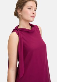 Robe fuchsia sans manches avec un décolleté drapé et un accent noué sur une épaule. Confectionnée dans un tissu léger et lisse.