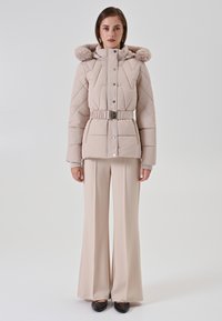 Vrouw in een beige gewatteerde jas met een met bont omzoomde capuchon, een ceintuur in de taille, brede beige broek en zwarte puntige schoenen, staande op een witte achtergrond.