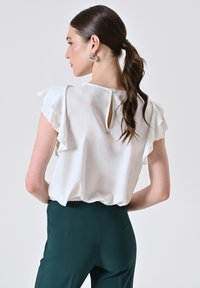 Donna con un ponytail scuro che indossa orecchini a cerchio argentati, una blusa bianca senza maniche con volant e pantaloni alta vita verde scuro, girata di lato.