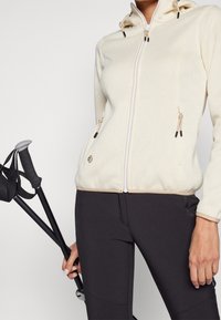 Person in einer cremefarbenen, strukturierten Zip-Jacke und schwarzen Hosen, der schwarze Wanderstöcke mit grauen Griffen hält.