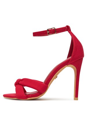 Sandalia roja de tacón alto con tira en el tobillo, tiras delanteras anudadas y tacón de aguja delgado sobre fondo blanco.