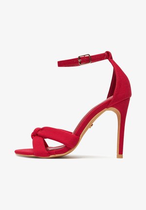 Sandalia roja de tacón alto con tira en el tobillo, tiras delanteras anudadas y tacón de aguja delgado sobre fondo blanco.