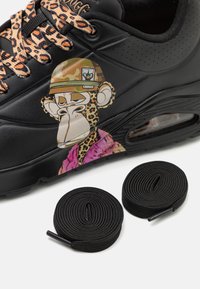 Schwarze Sneaker mit einem auffälligen Affengrafik, Leopardenmuster-Schnürsenkeln und einem rosa Akzent. Zwei gewickelte schwarze Schnürsenkel liegen in der Nähe.