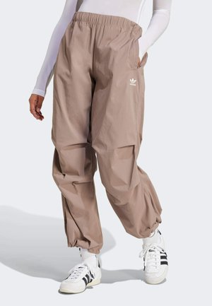 Personne portant un pantalon Adidas marron clair avec des poignets et une taille élastiques, un haut blanc à manches longues, et des baskets Adidas blanches avec des bandes noires.