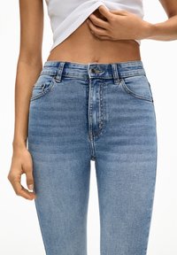 Jeans de mezclilla azul de cintura alta con un acabado desgastado, que cuentan con pasadores para cinturón y bolsillos frontales, creando una silueta ajustada.