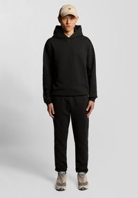 Felpa nera con cappuccio e pantaloni in tessuto morbido, abbinati a un berretto beige chiaro con un piccolo logo. Le scarpe sono grigie e beige.