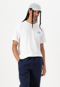 Weißes Baumwoll-T-Shirt mit einem türkisfarbenen Nike-Logo, kombiniert mit dunkelblauen Jogginghosen und einer hellgrauen Kappe, die geflochtene Haardetails aufweist.