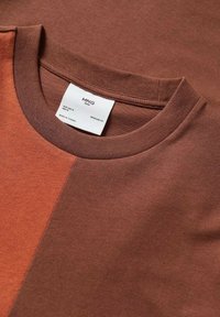 T-shirt en coton brun et couleur rouille avec col rond et étiquette blanche indiquant la taille moyenne et fabriqué en Turquie.