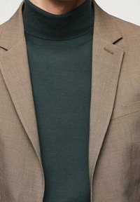 Beige bleiseri, jossa on teksturoitu kangas, yhdistetty tummanvihreän poolopaidan kanssa. Bleiserissä on hienovarainen ruutukuosi ja hartiaaksentti.