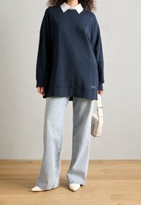 Sudadera de gran tamaño en color navy con un cuello blanco, combinada con jeans de pierna ancha en azul claro y zapatos blancos de punta, sosteniendo un bolso blanco.