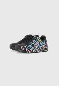 Baskets noires avec des motifs de coeurs pastel colorés, lacets noirs et semelles texturées, présentées de profil sur un fond blanc.