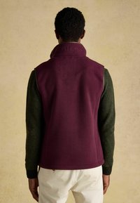 Fialová fleecová vesta s vysokým golierom a detailom loga; vrstvená nad tmavozelenou pletenou košeľou s dlhým rukávom; jednoduché biele nohavice.