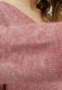 Rosa Strickpullover mit V-Ausschnitt, gerippter Textur und weichem, leicht flauschigem Stoff. Nahaufnahme des Ärmel-Details und der Farbvariationen.