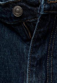 Gros plan d’un jean en denim bleu montrant un bouton métallique, une fermeture éclair et des coutures orange.