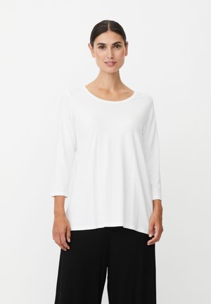 Vrouw staand, draagt een witte losse top met lange mouwen en zwarte wijde broekspijpen, tegen een effen lichte achtergrond.