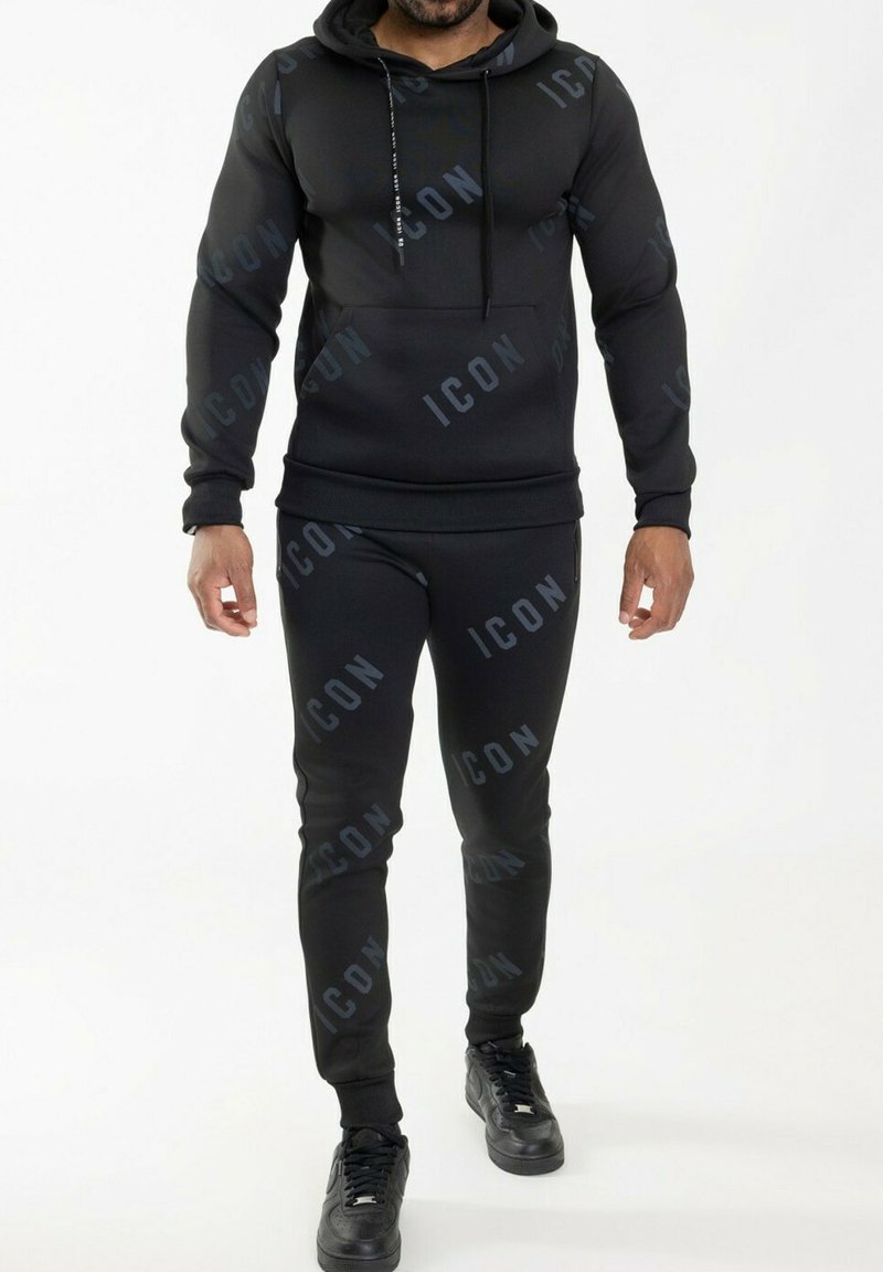 Ensemble de sweatpants et hoodie noirs avec un motif imprimé "ICON". Design ajusté, tissu doux, poignets côtelés et cordon de serrage. Sneakers noirs portés.